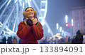 A young beautiful girl in a red coat walks in the evening at the New Year's Christmas fair against the backdrop of a Ferris wheel among lights and garlands and admires the decorations 133894091