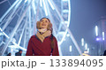 A young beautiful girl in a red coat walks in the evening at the New Year's Christmas fair against the backdrop of a Ferris wheel among lights and garlands and admires the decorations 133894095