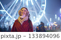 A young beautiful girl in a red coat walks in the evening at the New Year's Christmas fair against the backdrop of a Ferris wheel among lights and garlands and admires the decorations 133894096