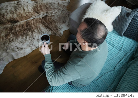 Caucasian man sitting with a cup in hand on sofa indoors, enjoying a calm moment of rest and comfort in a cozy home space. Home Leisure Activities 133894145
