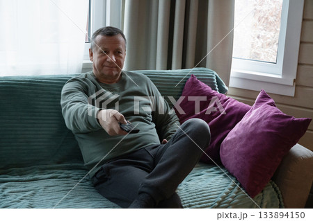 Caucasian man sitting on sofa and watching TV with remote control, enjoying a quiet and comfortable indoor moment at home. Home Leisure Activities Caucasian man sitting on sofa and watching TV with remote control, enjoying a quiet and comfortable indoor moment at home. Home Leisure Activities 133894150