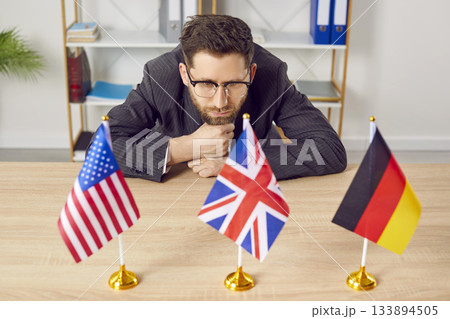 Pensive businessman sitting at desk looking at flags of different countries 133894505