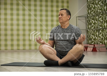 Practicing seated meditation senior caucasian man sitting in lotus pose in indoor fitness studio calming mind and restoring focus after long workday 133895789