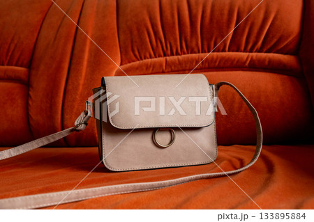 Stylish beige crossbody handbag resting on a vibrant orange sofa. eco-suede bag with a long strap, shoulder bag Stylish beige crossbody handbag resting on a vibrant orange sofa. eco-suede bag with a long strap, shoulder bag 133895884