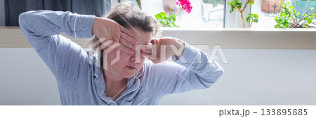A 50-year-old woman appears tired and distressed as she sits up in bed. She rubs her eyes, indicating a lack of sleep, while surrounded by soft bedding in a bright room, banner A 50-year-old woman appears tired and distressed as she sits up in bed. She rubs her eyes, indicating a lack of sleep, while surrounded by soft bedding in a bright room, banner 133895885