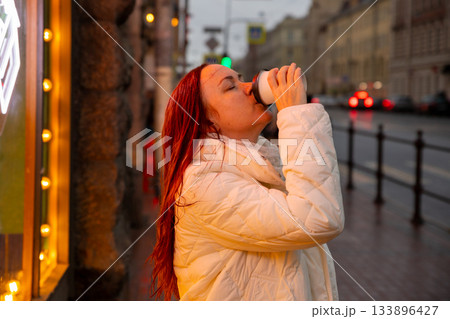 middle aged Caucasian woman drinks coffee on street in metropolis in evening in rain. 133896427