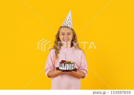 Smiling preteen girl in party hat holding birthday cake with lit candle Smiling preteen girl in party hat holding birthday cake with lit candle 133897659