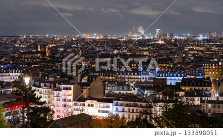 Cityscape of Paris, France glows at night seen from Montmartre hill. Panoramic view features illuminated rooftops, streetlights, and urban horizon under cloudy sky in Ile-de-France region Cityscape of Paris, France glows at night seen from Montmartre hill. Panoramic view features illuminated rooftops, streetlights, and urban horizon under cloudy sky in Ile-de-France region 133898051