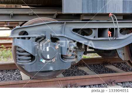 Metal freight wagon bogie with heavy steel springs and worn wheel surface stands on rusty tracks under daylight. Industrial structure shows suspension, bearings and connections with visible textures Metal freight wagon bogie with heavy steel springs and worn wheel surface stands on rusty tracks under daylight. Industrial structure shows suspension, bearings and connections with visible textures 133904051