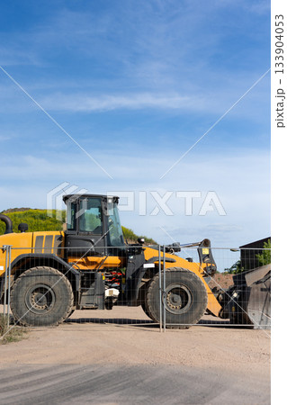 Bright yellow wheel loader stands behind metal fence on dusty construction ground. Sunlight highlights large tires, cabin and bucket while green hill and buildings form calm industrial backdrop 133904053