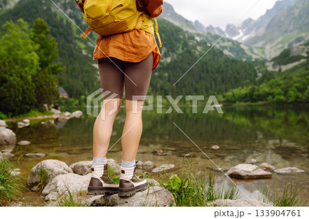 Travel Slovakia, Europe. Tourist with a yellow backpack stands against the backdrop of alpine lake. Travel Slovakia, Europe. Tourist with a yellow backpack stands against the backdrop of alpine lake. 133904761