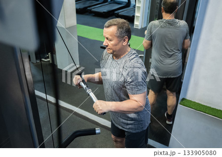 Senior man presses cable handle downward while training triceps in gym, maintaining controlled movement and strength focus. Working Out 133905080