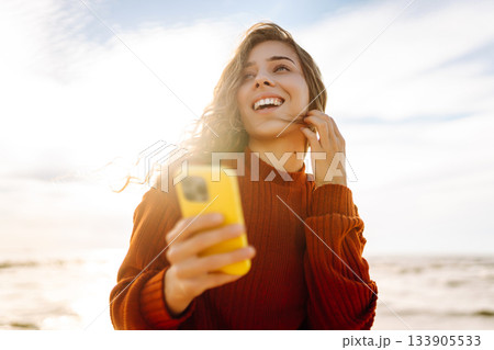 Young woman using mobile phone at beach. Cold weather. Autumn. Blogging, communication Young woman using mobile phone at beach. Cold weather. Autumn. Blogging, communication 133905533