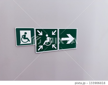 Green sign indicating gathering point for people with disabilities. This point has accessible path for wheelchair users. Arrows point to disabled symbol on wall. Concept of accessible environment Green sign indicating gathering point for people with disabilities. This point has accessible path for wheelchair users. Arrows point to disabled symbol on wall. Concept of accessible environment 133906818