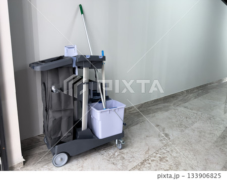 Cleaning cart with large trash bag stands in hallway. Mobile cleaning trolley with professional equipment. Mop, bucket, and trash can are on wheeled cart Cleaning cart with large trash bag stands in hallway. Mobile cleaning trolley with professional equipment. Mop, bucket, and trash can are on wheeled cart 133906825