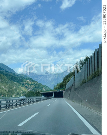Mountain highway leads through lush landscapes to a distant tunnel under a partly cloudy sky. Green hills and majestic peaks create a serene road trip experience full of natural beauty. Mountain highway leads through lush landscapes to a distant tunnel under a partly cloudy sky. Green hills and majestic peaks create a serene road trip experience full of natural beauty. 133907023