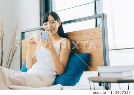 ベッドでコーヒーを飲みながらスマホを見る若い女性 133907537