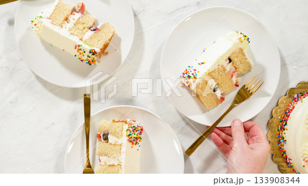Two slices of vanilla cake with rainbow sprinkles and white frosting are served on round white plates. The scene is styled with gold forks on a marble surface, evoking a festive and cheerful mood. 133908344