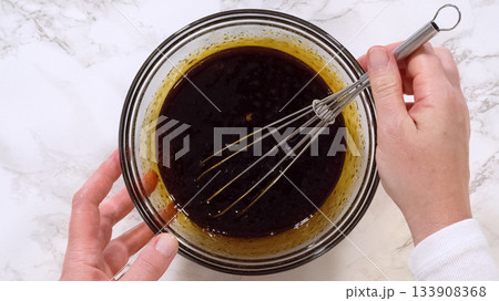 Hands whisk a glossy dark liquid mixture inside a glass bowl placed on a marble countertop. The mixture appears rich and smooth, representing a key step in preparing cookie dough. Bright light 133908368
