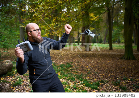 A man with a beard operates a drone while celebrating its flight. Colorful leaves surround him in a serene park during autumn. Joy fills the air as he engages in this activity. A man with a beard operates a drone while celebrating its flight. Colorful leaves surround him in a serene park during autumn. Joy fills the air as he engages in this activity. 133909402