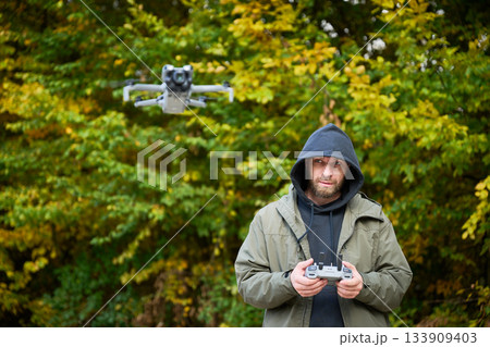 A man controls a drone with concentration amidst a colorful forest. Yellow and green leaves surround him, creating a beautiful backdrop during a calm autumn day. 133909403