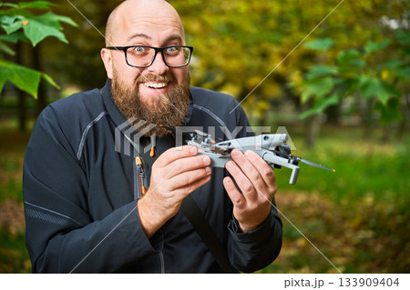 A joyful man showcases his new drone against a backdrop of lush green trees in a lively park. His excitement is evident as he holds the device with care. 133909404