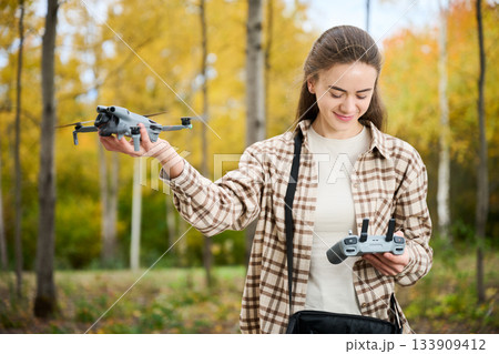 In a lively autumn forest, a woman holds her drone confidently while preparing to launch it with a remote control. Surrounding trees display beautiful fall colors. In a lively autumn forest, a woman holds her drone confidently while preparing to launch it with a remote control. Surrounding trees display beautiful fall colors. 133909412