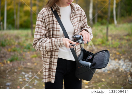 In a serene location filled with autumn leaves, a young woman adjusts her camera while looking focused and ready to capture the beauty around her. In a serene location filled with autumn leaves, a young woman adjusts her camera while looking focused and ready to capture the beauty around her. 133909414