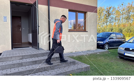 A man in a gray and orange jacket walks down concrete steps outside a building, carrying a black duffel bag toward parked cars on a grassy area under a blue sky 133909567