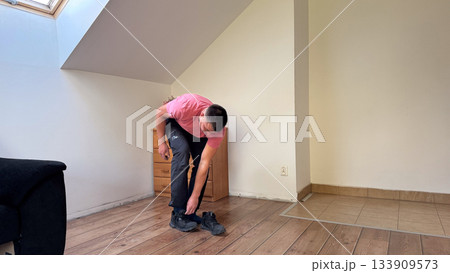 A man in a pink t-shirt and black pants leans forward to put on black sneakers in a simple attic room with wooden and tiled flooring 133909573