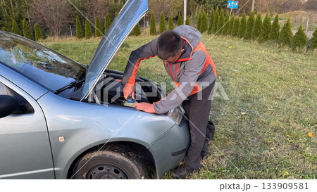 A man in a gray and orange jacket tightens the cap on a fluid reservoir under the hood of a silver car during roadside maintenance in a grassy rural area A man in a gray and orange jacket tightens the cap on a fluid reservoir under the hood of a silver car during roadside maintenance in a grassy rural area 133909581
