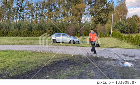 A man in a gray and orange jacket walks across a paved path and grassy area carrying a tool bag, approaching a silver car parked on grass for roadside repair 133909587