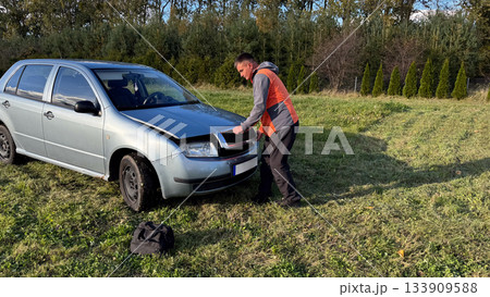 A man in a gray and orange jacket opens the hood of a silver car parked on grass, preparing to inspect and repair the engine during roadside maintenance in a rural area 133909588