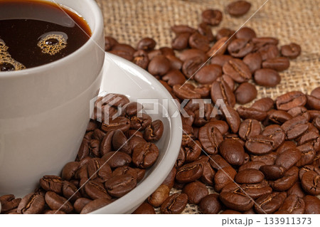 A beautiful cup of coffee surrounded by scattered coffee beans on a rustic burlap background A beautiful cup of coffee surrounded by scattered coffee beans on a rustic burlap background 133911373