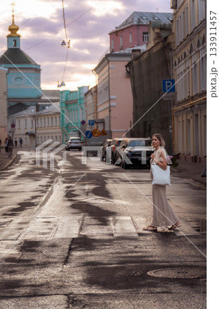 A woman crosses a quiet city street at sunset with a white tote bag and pink hydrangea, warm light reflecting on the wet pavement 133911457