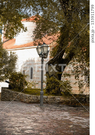 View of a historic church next to an olive tree and a traditional street lantern in the Old Town of Budva, Montenegro. Showcases medieval architecture, stone walls, narrow streets, and Mediterranean View of a historic church next to an olive tree and a traditional street lantern in the Old Town of Budva, Montenegro. Showcases medieval architecture, stone walls, narrow streets, and Mediterranean 133911799