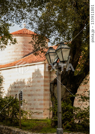 Charming historic church located next to an olive tree and a classic street lantern in the Old Town of Budva, Montenegro. Highlights medieval architecture, stone textures, narrow streets, and 133911801