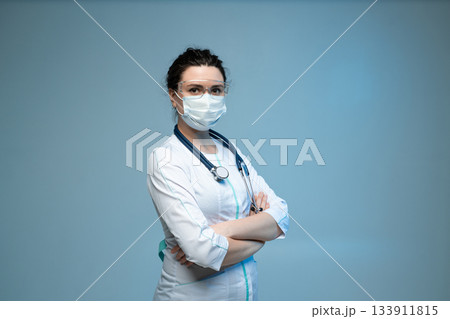 Medical worker in mask and protective glasses standing confidently with folded arms on blue background Medical worker in mask and protective glasses standing confidently with folded arms on blue background 133911815