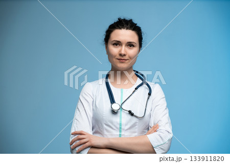 Female doctor with stethoscope posing confidently against blue background in professional uniform Female doctor with stethoscope posing confidently against blue background in professional uniform 133911820