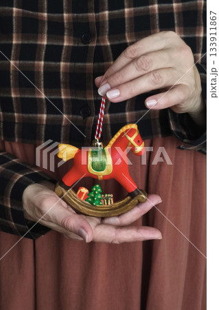 Woman in plaid and terracotta dress holds red rocking horse ornament with tree and gifts, cozy rustic holiday mood 133911867