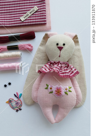 Handmade soft rabbit doll in pastel tones surrounded by pink craft supplies, delicate feminine vibe 133911870