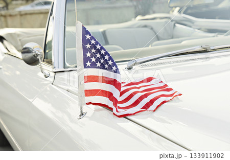 Small American flag mounted on antenna of white vintage convertible, stars and stripes draped over hood in bright daylight 133911902