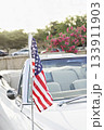 Close view of American flag on antenna of white convertible with chrome and light leather interior, soft flower bokeh in daylight 133911903