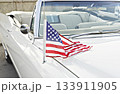 Small American flag mounted on antenna of white classic convertible, stars and stripes draped across hood in bright daylight, patriotic summer vibe 133911905