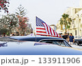Small American flag on car roof at city street event, people and palm trees in soft background, bright summer vibe 133911906
