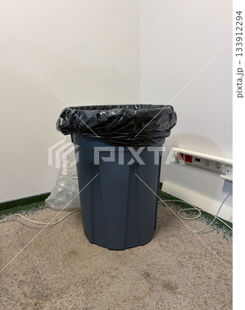 A gray plastic trash bin with a black liner stands against a white wall near electrical sockets and loose cables on a rough floor A gray plastic trash bin with a black liner stands against a white wall near electrical sockets and loose cables on a rough floor 133912294