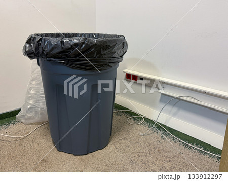A gray plastic trash bin with a black liner stands against a white wall near electrical sockets and loose cables on a rough floor 133912297