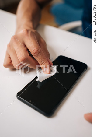 Vertical cropped shot of male hand cleaning screen of smartphone with disinfectant wipe, promoting hygiene and effectively eliminating germs and bacteria for better health, sitting at white home desk Vertical cropped shot of male hand cleaning screen of smartphone with disinfectant wipe, promoting hygiene and effectively eliminating germs and bacteria for better health, sitting at white home desk 133912768