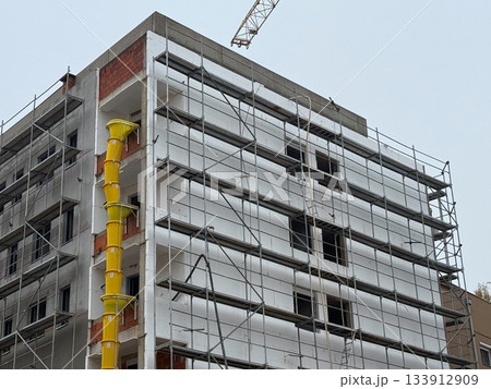 Building under construction with scaffolding and yellow chute. Urban development, engineering, and architectural process of modern residential growth. 133912909