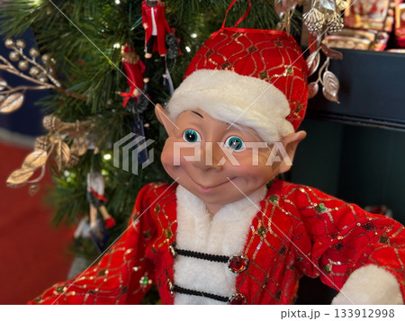 Cheerful elf toy in festive red outfit near Christmas tree. Holiday spirit, winter magic and childhood nostalgia, celebration atmosphere and tradition warmth. 133912998